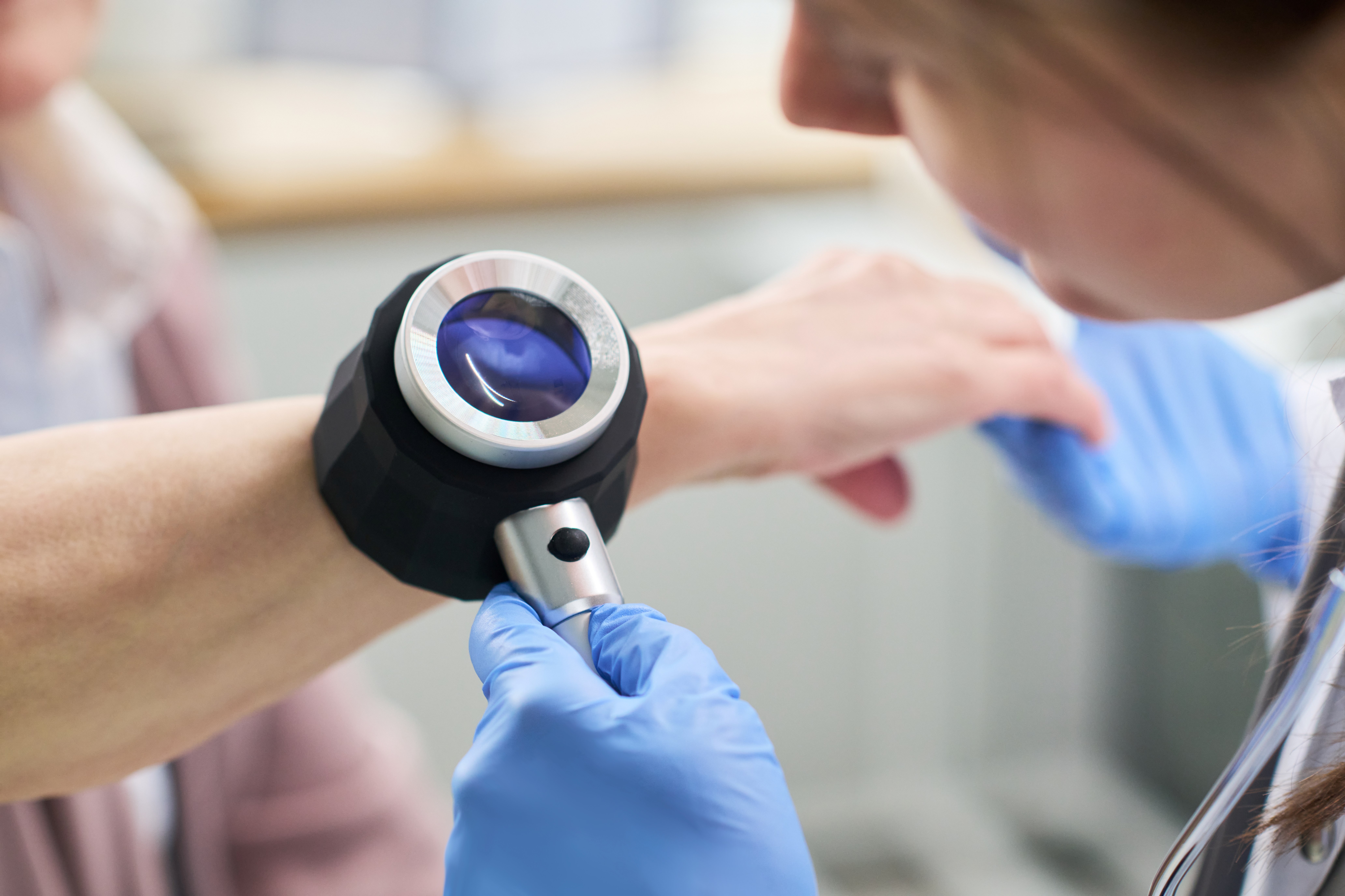 Adult woman examining forearm of senior woman using magnifying device during dermatological checkup, gloved hands holding instrument close to skin